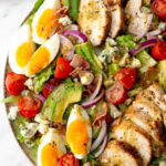 A salad bowl with sliced grilled chicken, avocado, tomato, red onion, hard-boiled eggs, bacon, blue cheese, and lettuce, topped with vinaigrette dressing.
