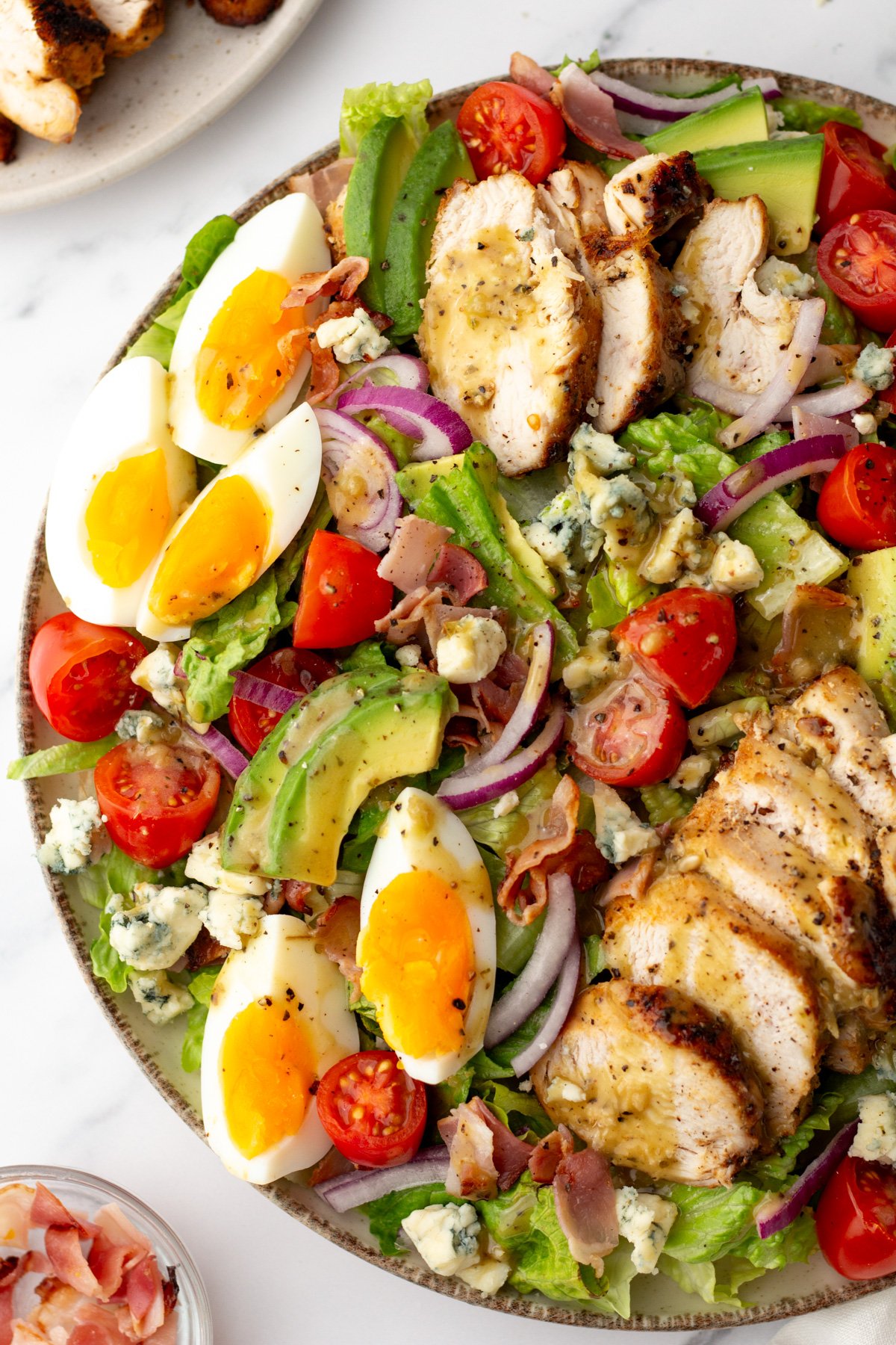 A salad bowl with sliced grilled chicken, avocado, tomato, red onion, hard-boiled eggs, bacon, blue cheese, and lettuce, topped with vinaigrette dressing.