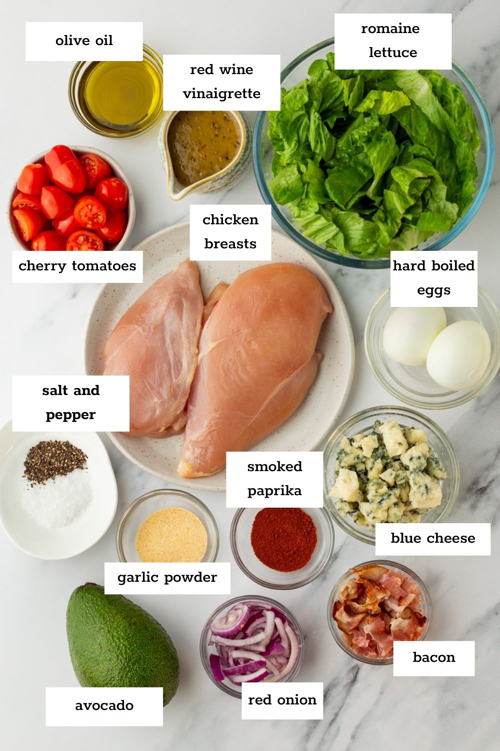 Top-down view of ingredients for a salad, including chicken breasts, lettuce, hard-boiled eggs, cherry tomatoes, bacon, blue cheese, avocado, red onion, olive oil, and seasonings.