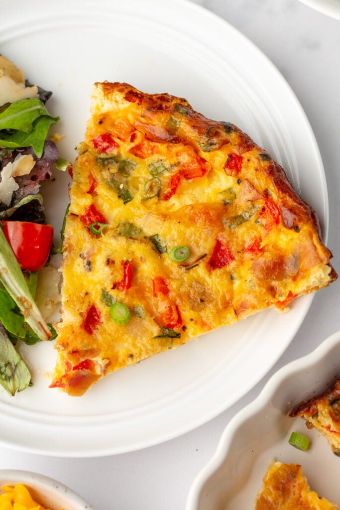 A slice of vegetable frittata with visible red peppers, green onions, and herbs on a white plate next to a mixed green salad.