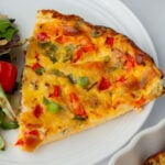 A slice of vegetable quiche with visible green onions, red peppers, and cheese on a white plate next to a side salad.