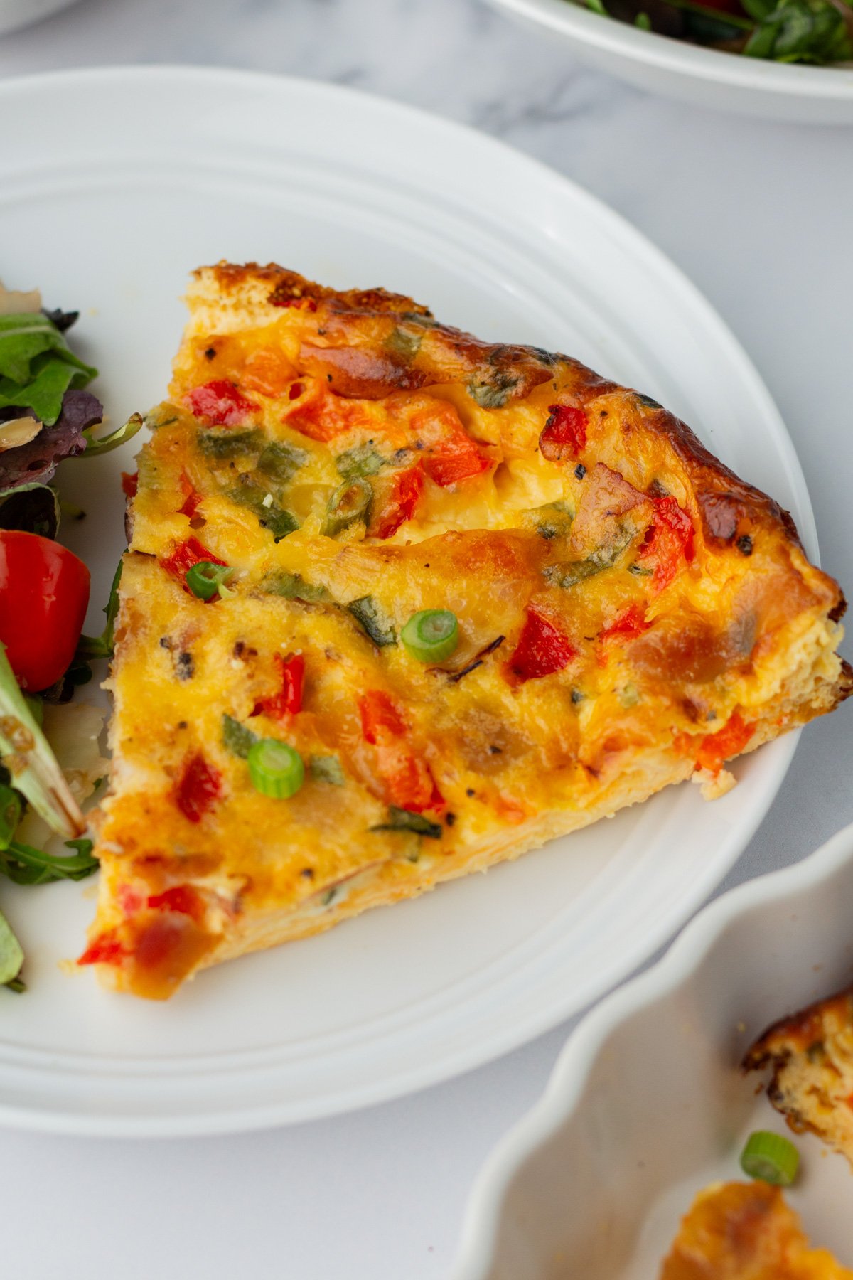 A slice of vegetable quiche with visible green onions, red peppers, and cheese on a white plate next to a side salad.