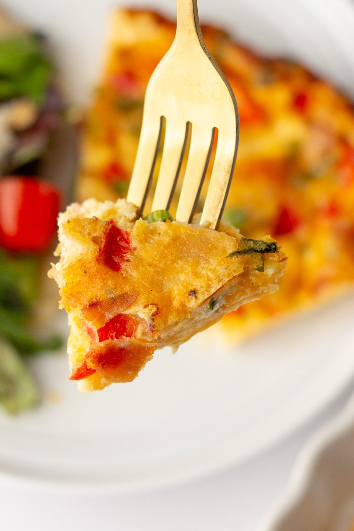A fork holds a bite of vegetable frittata with visible red bell peppers and green herbs; a partial salad and plate are in the background.