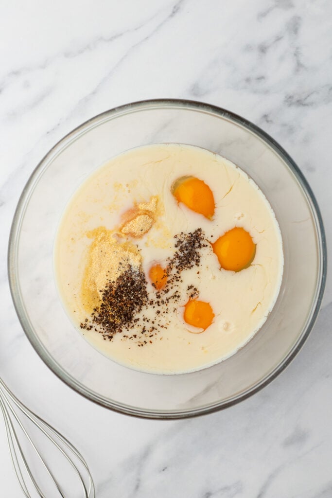 A glass mixing bowl containing eggs, cream, ground black pepper, and seasoning on a marble surface, with a metal whisk nearby.