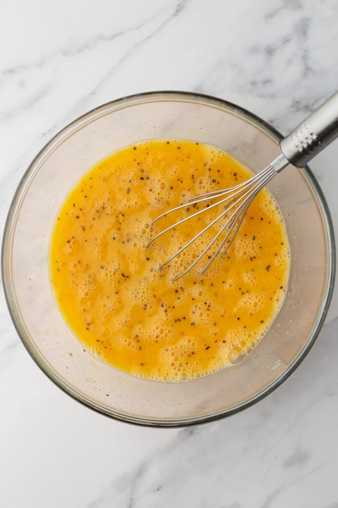 A glass bowl containing beaten eggs with black pepper, and a metal whisk resting inside, on a marble surface.