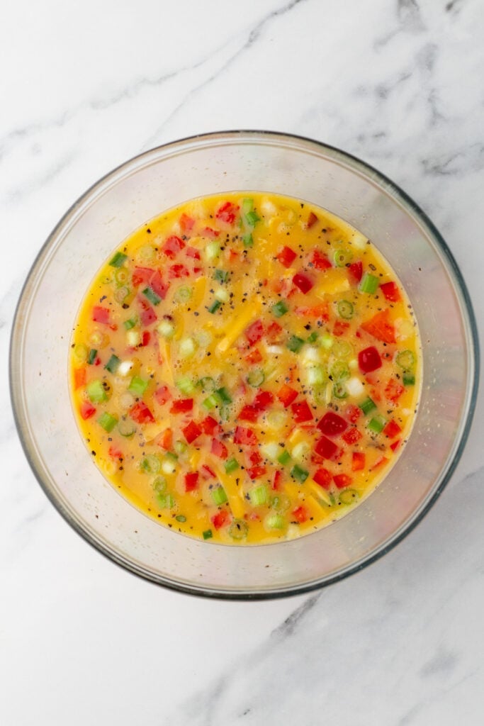 A glass bowl filled with a mixed egg mixture containing chopped red and green bell peppers, green onions, and black pepper, on a marble surface.