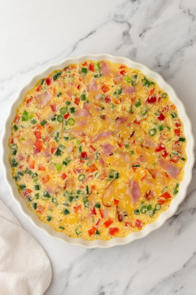 A round baking dish filled with an uncooked egg mixture containing chopped ham, green onions, and red bell peppers on a marble surface.