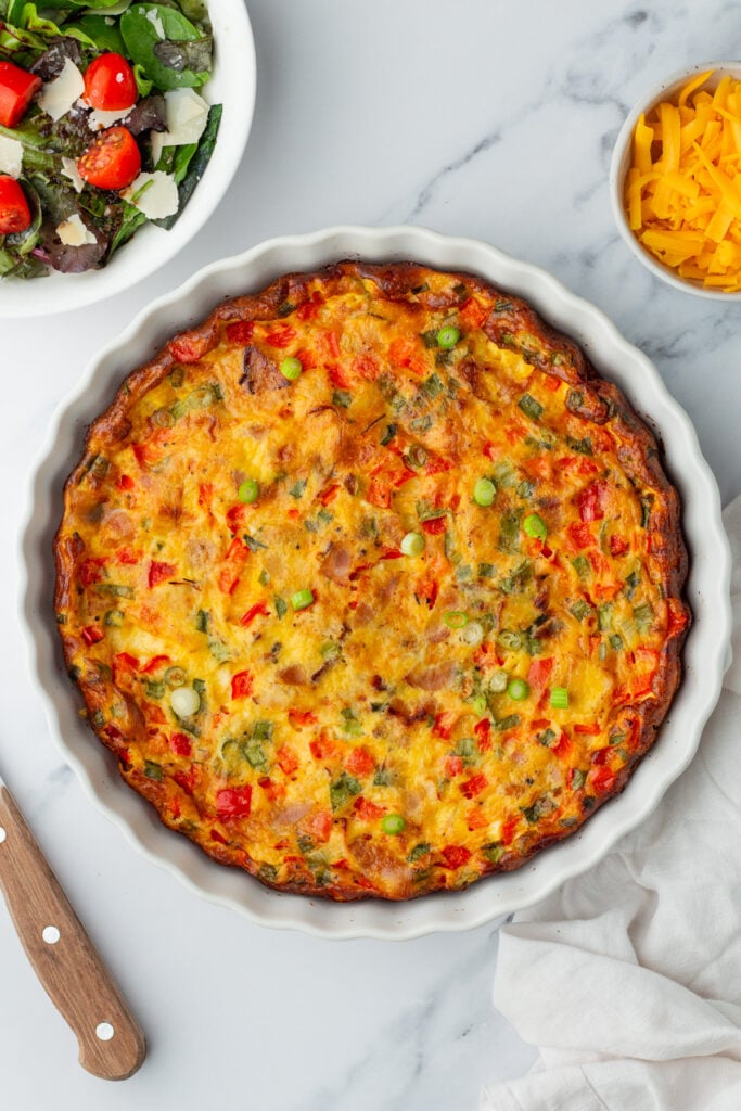 A baked vegetable quiche in a round dish, surrounded by a salad, shredded cheddar cheese, and a wooden-handled knife on a marble surface.