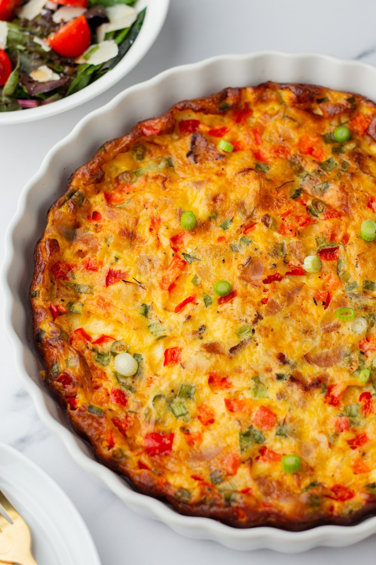A baked quiche with a golden-brown crust, topped with chopped green onions and red bell peppers, in a white dish next to a salad.