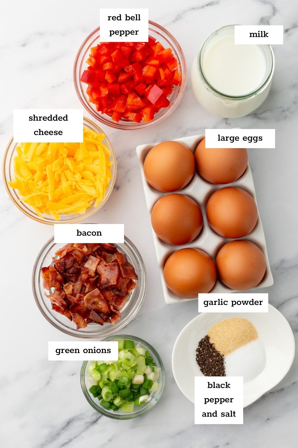 Various labeled ingredients including red bell pepper, milk, shredded cheese, large eggs, bacon, green onions, garlic powder, black pepper, and salt arranged on a white surface.