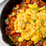 Scrambled eggs topped with black pepper and chopped green onions served over a hash of sweet potatoes, bacon, and vegetables in a black skillet.