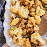 A white platter with air-fryer cauliflower florets, some garnished with herbs, and a wooden serving spoon on the side.