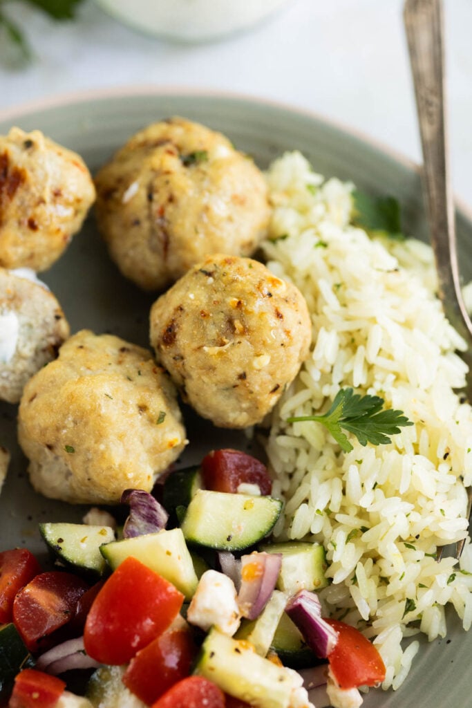 A plate with Greek chicken meatballs, a serving of rice, and a salad made of cucumbers, tomatoes, red onions, and feta cheese.
