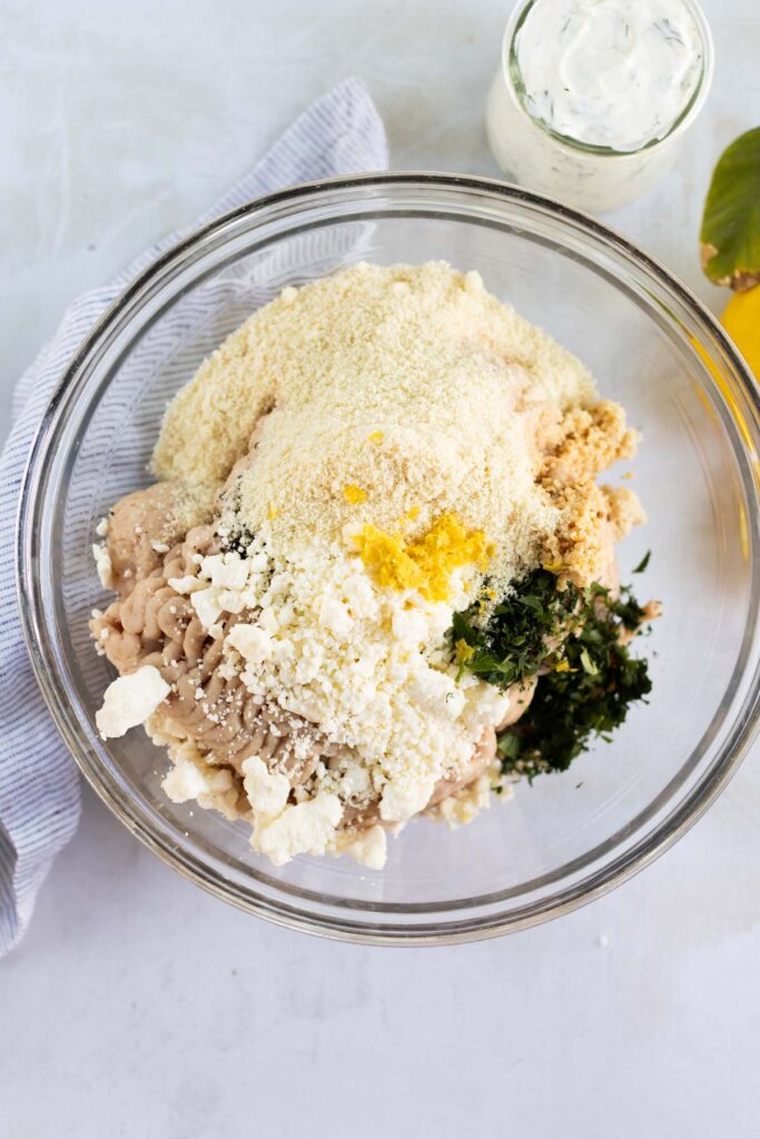 A glass bowl containing ground chicken, almond flour, feta cheese, herbs, and lemon zest, with a jar of creamy sauce nearby on a white surface.
