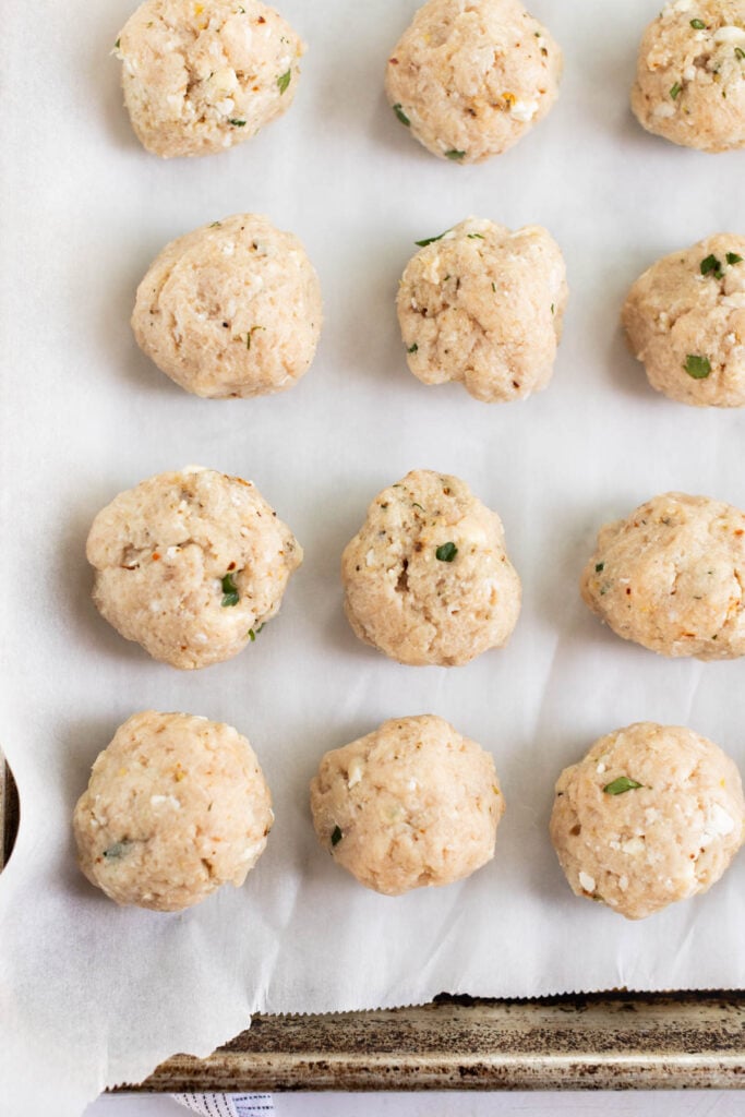Raw chicken meatballs mixed with herbs and seasonings are arranged in neat rows on a parchment-lined baking sheet.