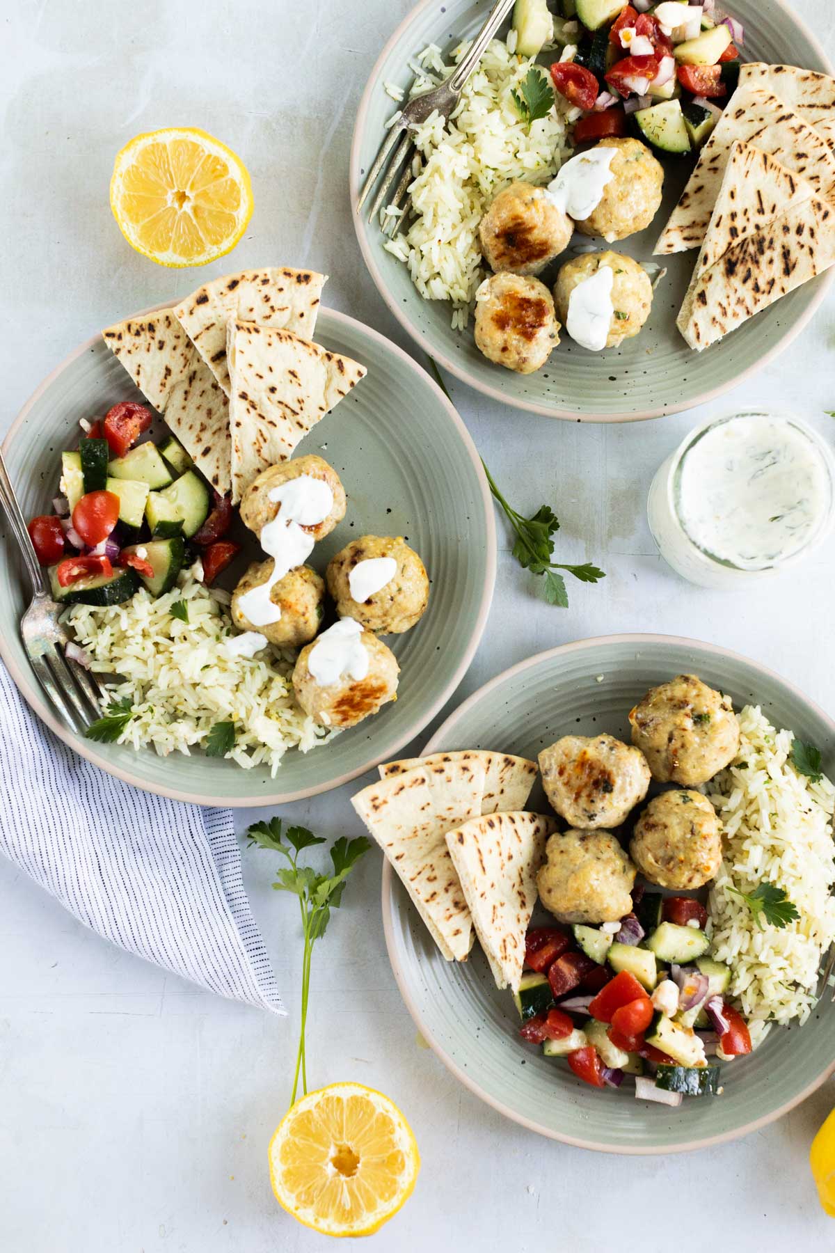 Three plates with rice, pita bread, salad with cucumbers and tomatoes, Greek chicken meatballs topped with yogurt sauce, and lemon halves on the side.