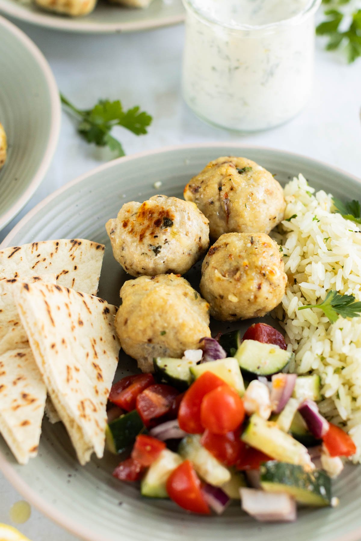 A plate with Greek chicken meatballs, flatbread slices, white rice with herbs, and a colorful chopped vegetable salad.