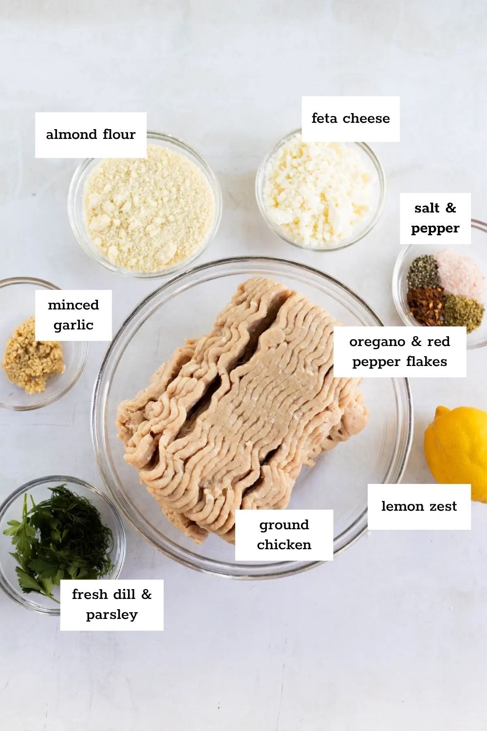 A top-down view of labeled ingredients in bowls for Greek chicken meatballs, including ground chicken, almond flour, feta cheese, seasonings, minced garlic, lemon zest, and fresh herbs.