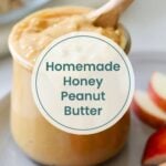 A jar of creamy homemade honey peanut butter with a wooden spoon, placed next to sliced apples on a plate—perfect as a snack or paired with high protein chicken salad for a satisfying meal.