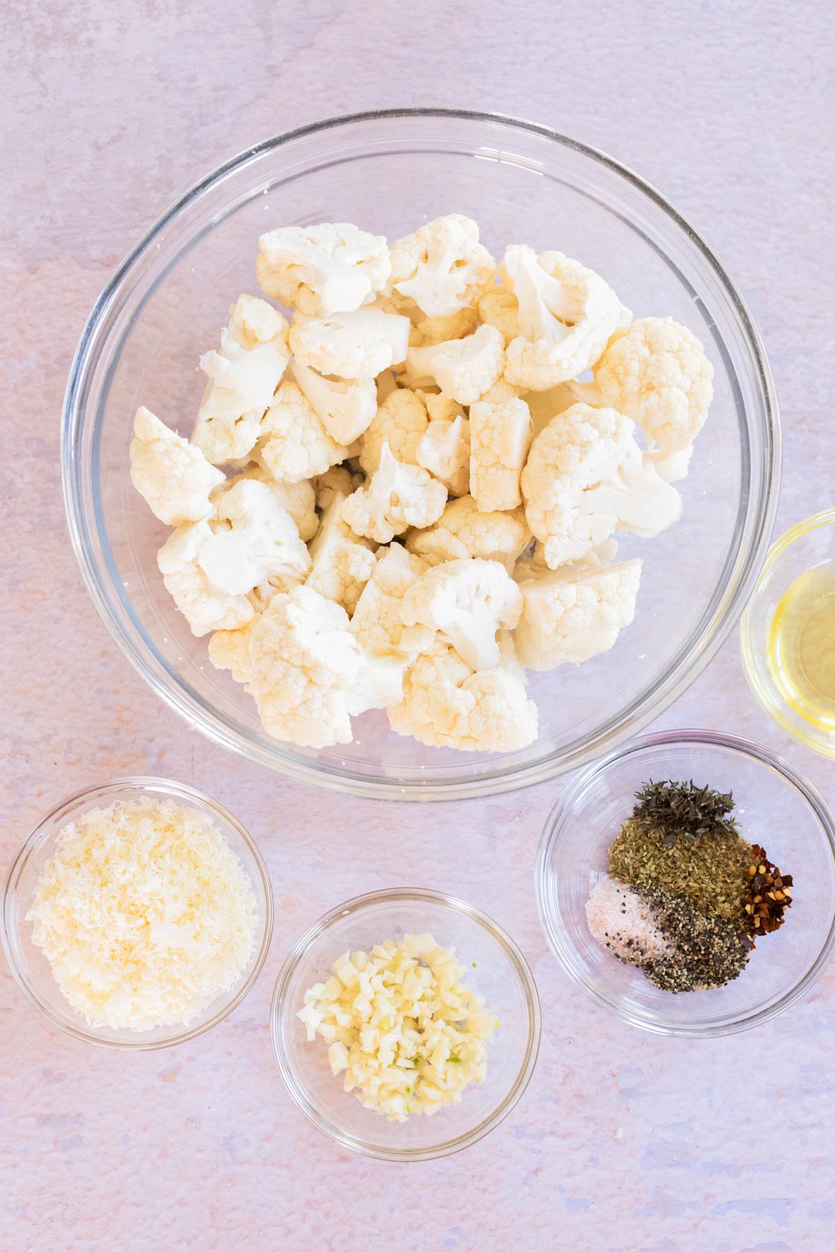 A bowl of cauliflower florets surrounded by small bowls of grated cheese, chopped garlic, mixed dried herbs and spices, and oil on a light surface.