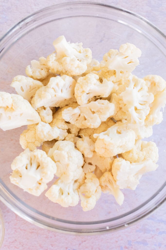 A clear glass bowl filled with raw cauliflower florets, viewed from above on a light surface.