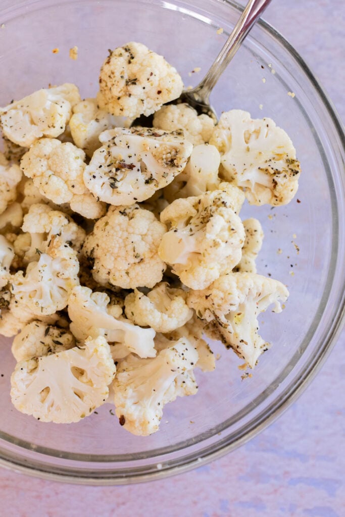 A glass bowl filled with raw cauliflower florets seasoned with herbs and spices, with a metal spoon inside.