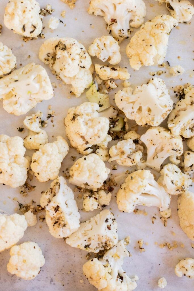 Raw cauliflower florets coated with herbs and spices are scattered on a baking sheet lined with parchment paper.