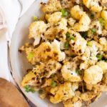 A plate of roasted cauliflower florets garnished with chopped green onions, next to a wooden spoon and a white cloth napkin.