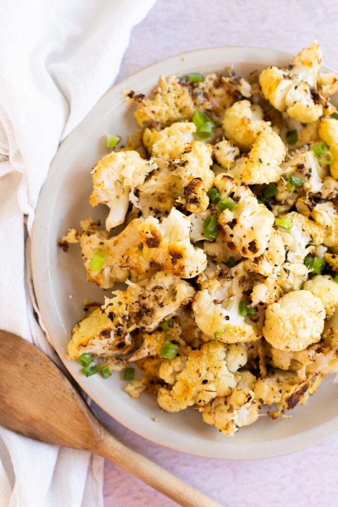 A plate of roasted cauliflower florets garnished with chopped green onions, next to a wooden spoon and a white cloth napkin.
