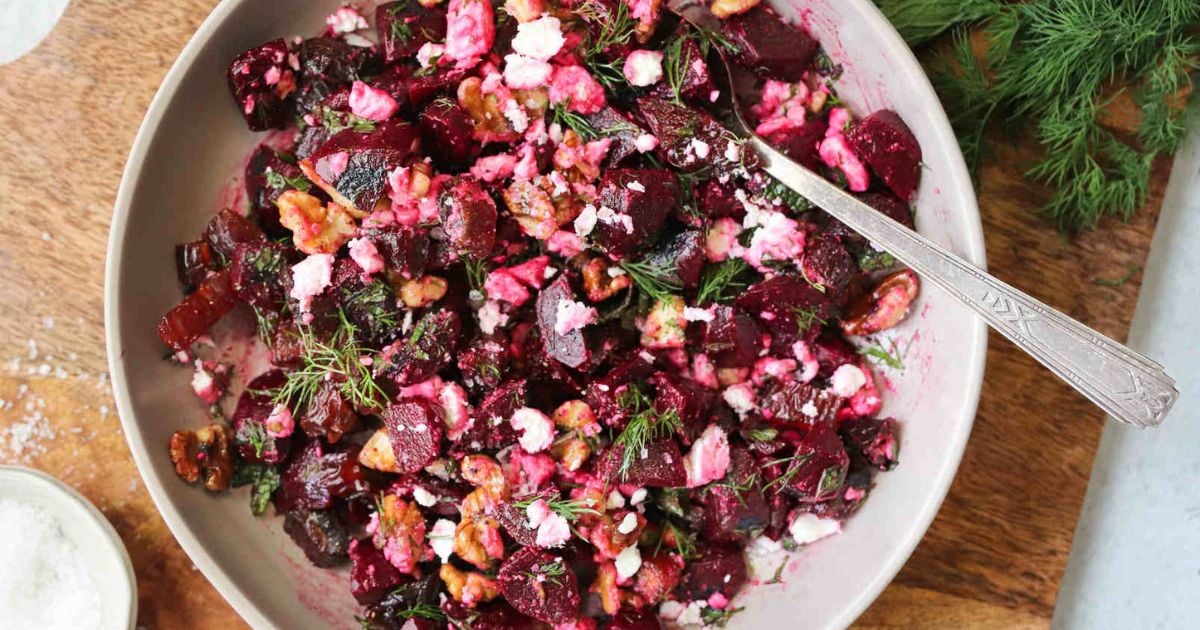 A bowl of beet salad with crumbled feta, walnuts, and fresh dill, mixed together with a spoon on a wooden surface.