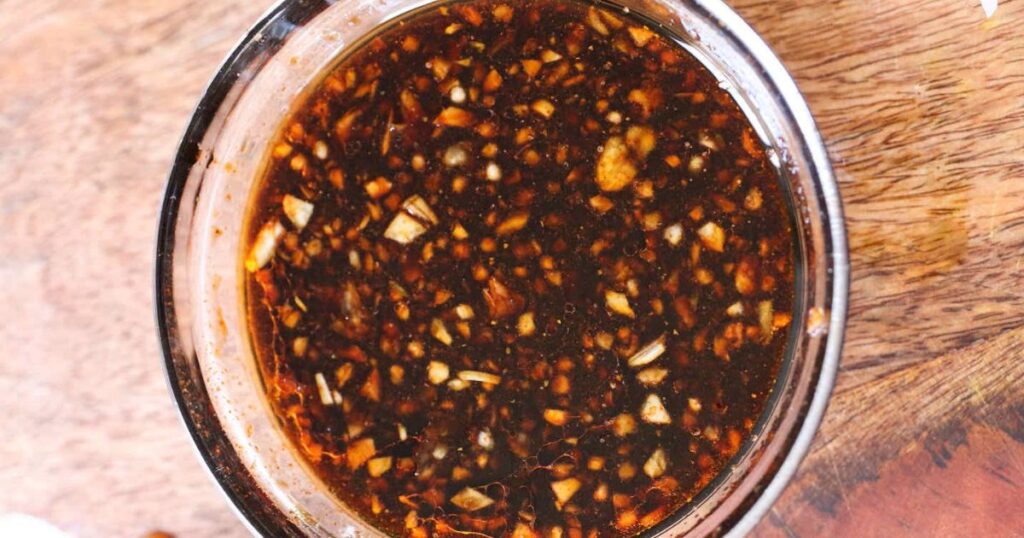 A close-up view of a glass bowl filled with a chunky, dark-colored sauce containing chopped garlic and spices, placed on a wooden surface.