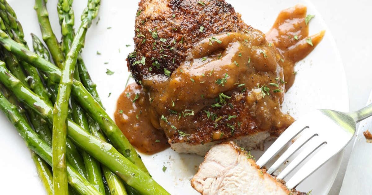A cooked pork chop topped with brown sauce, served with sautéed asparagus on a white plate, with a fork cutting into the meat.