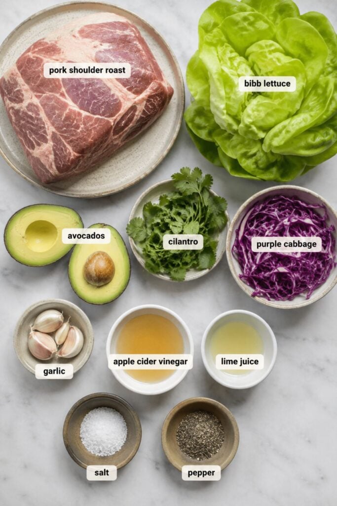 Ingredients for pork lettuce wraps arranged on a surface: pork shoulder roast, bibb lettuce, avocado, cilantro, purple cabbage, garlic, apple cider vinegar, lime juice, salt, and pepper.