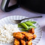 A plate with white rice, sugar snap peas, and air-fryer salmon bites, with a fork, next to an air fryer containing more chicken.