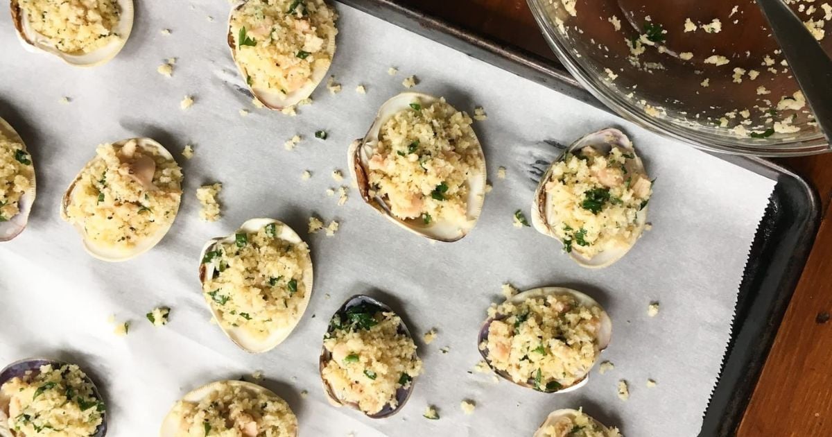 Stuffed clams topped with breadcrumbs and herbs are arranged on a parchment-lined baking sheet, ready to be cooked. A mixing bowl with extra stuffing is visible in the corner.