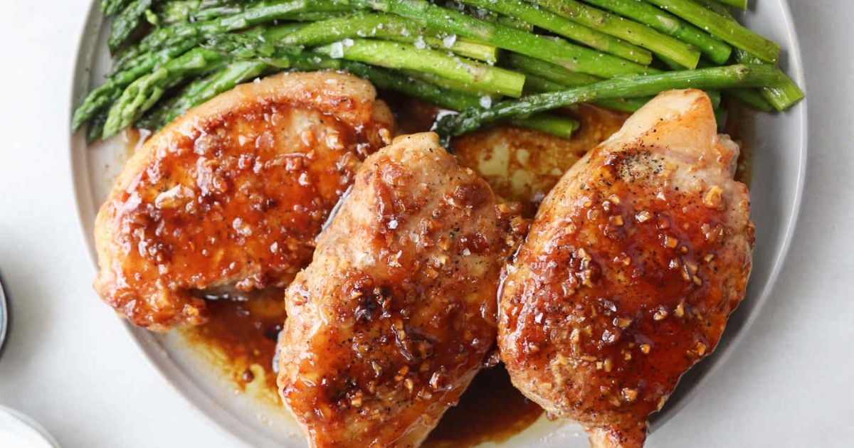 Three cooked chicken breasts with a glazed sauce are served on a plate alongside a portion of seasoned asparagus spears.