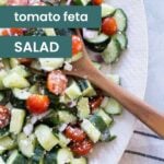 A wooden spoon rests in a bowl of cucumber tomato feta salad with red onion, served on a white plate, accompanied by text overlay describing the fresh flavors.