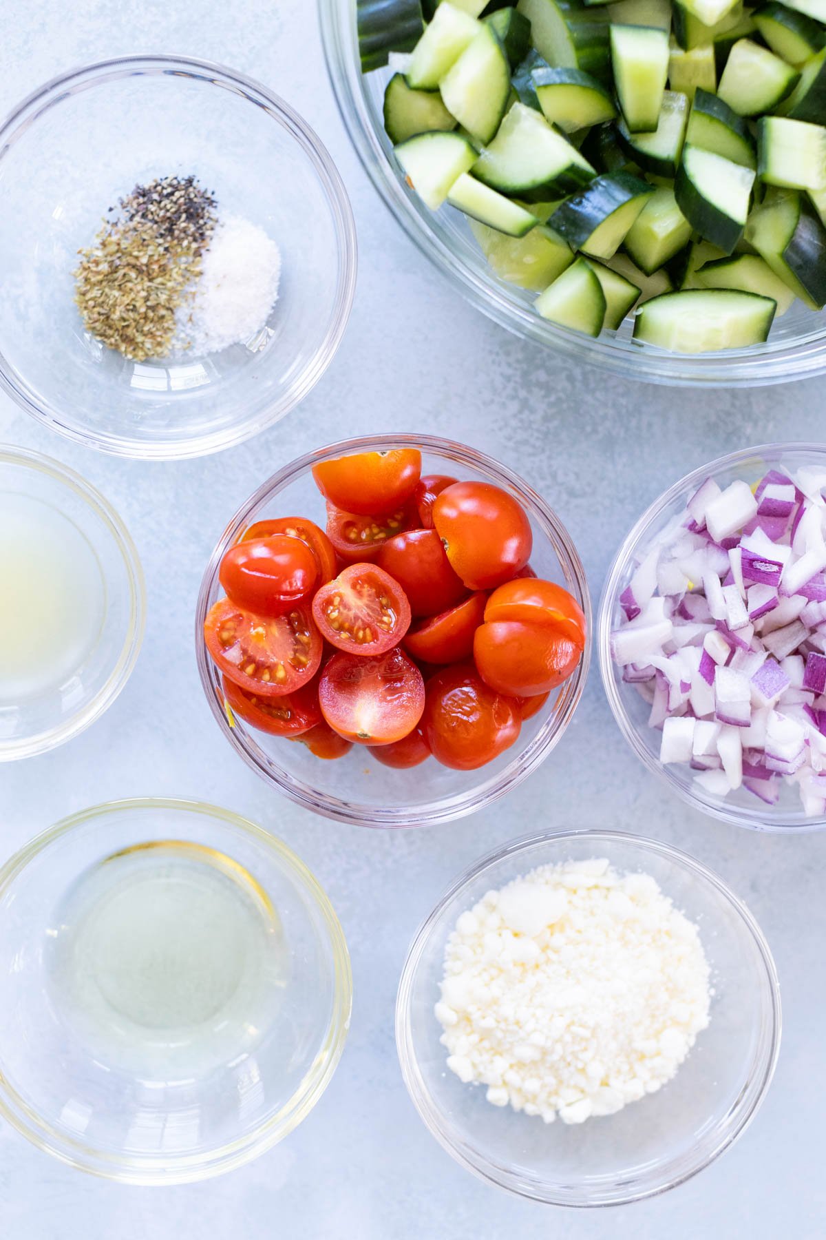 Bowls containing chopped cucumber, halved cherry tomatoes, diced red onion, crumbled feta cheese, dried herbs with salt, olive oil, and lemon juice arranged on a light surface.