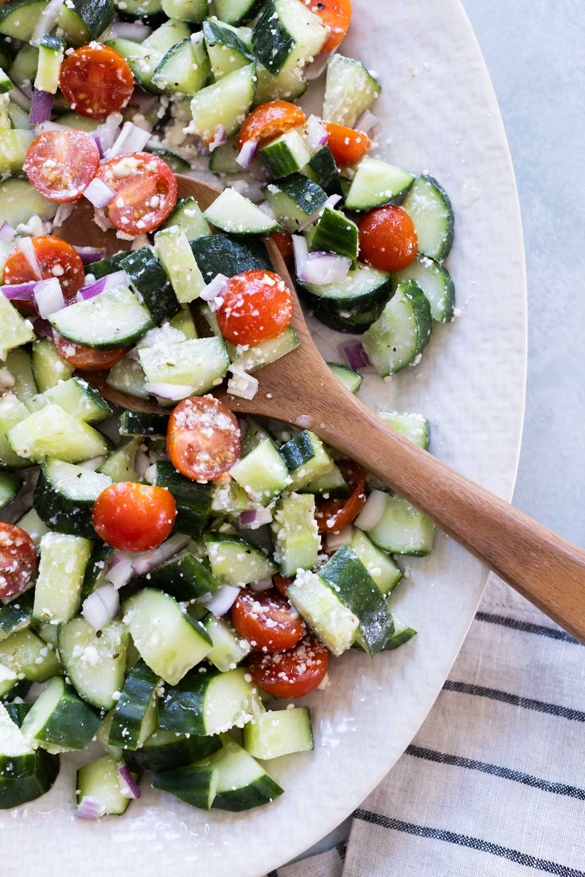 A wooden spoon rests in a bowl of chopped cucumber, cherry tomatoes, red onion, and crumbled cheese salad on a white plate.