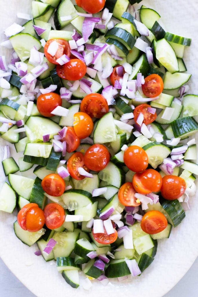 Sliced cucumbers, cherry tomatoes, and chopped red onions arranged on a white surface.