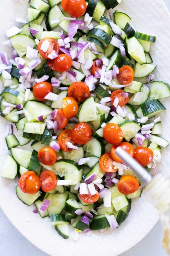A salad with sliced cucumbers, cherry tomatoes, and chopped red onions on a white platter, with a dressing being poured from a jar.