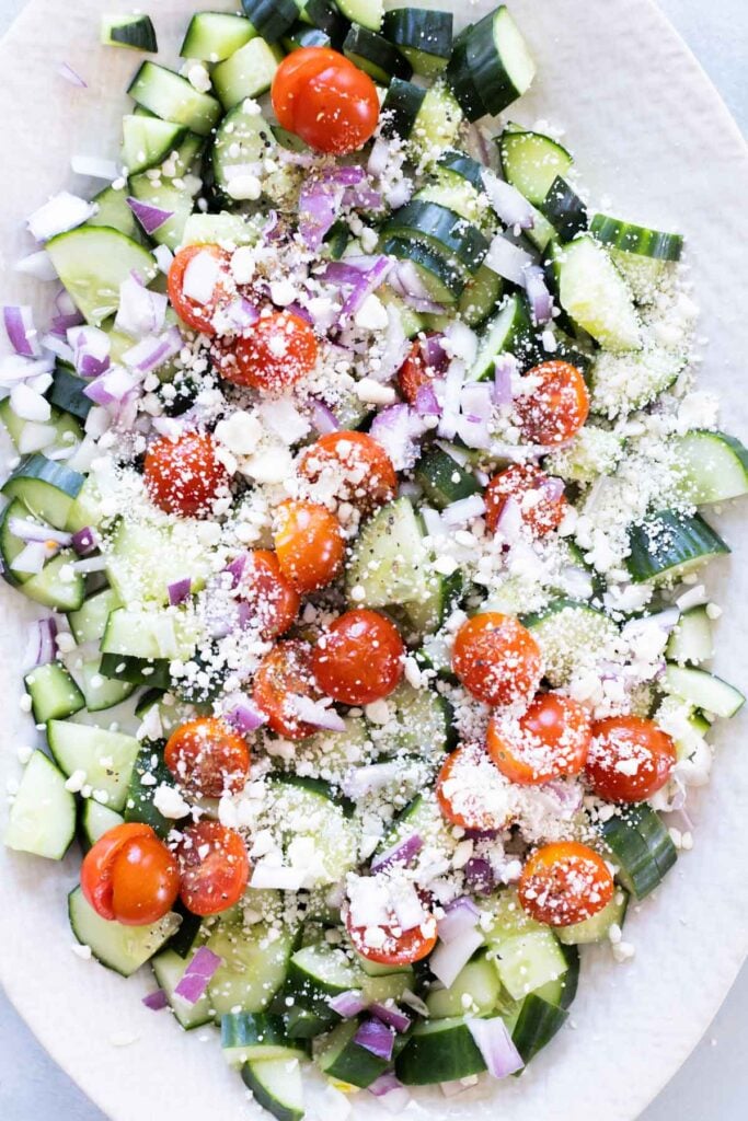 Chopped cucumber, cherry tomatoes, red onion, and crumbled feta cheese arranged on a white plate.
