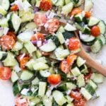 Chopped cucumber, cherry tomatoes, red onion, and feta cheese mixed in a bowl with a wooden spoon.