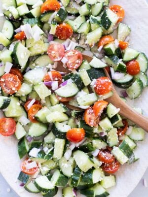Chopped cucumber, cherry tomatoes, red onion, and feta cheese mixed in a bowl with a wooden spoon.
