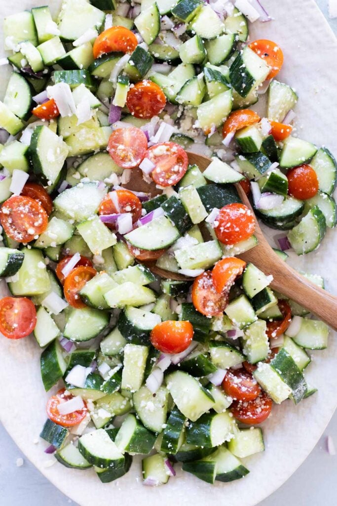Chopped cucumber, cherry tomatoes, red onion, and feta cheese mixed in a bowl with a wooden spoon.