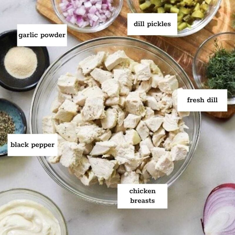 A marble counter displays bowls of chopped chicken breasts, red onion, dill pickles, fresh dill, mayo, garlic powder, and black pepper—everything you need to make a flavorful dill pickle chicken salad. Each ingredient is labeled with text.