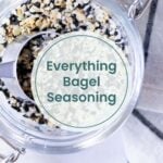 A jar of everything bagel seasoning with a metal scoop sits beside a striped cloth, viewed from above.