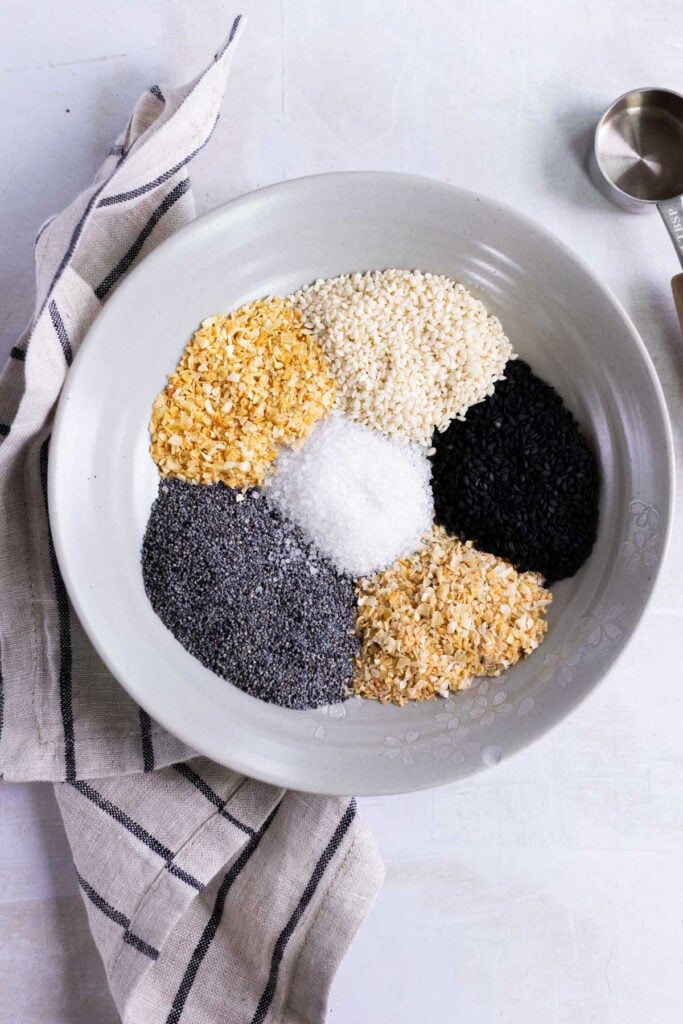 A white bowl containing separate piles of dried minced onion, white sesame seeds, black sesame seeds, poppy seeds, dried garlic, and salt, with a striped cloth beside it.