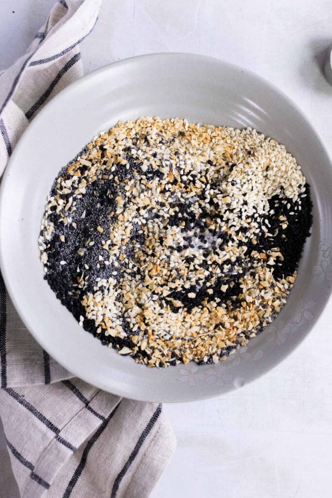 A white bowl containing a mixture of black and white sesame seeds, dried minced garlic, and dried onion, placed on a light surface with a striped cloth beside it.