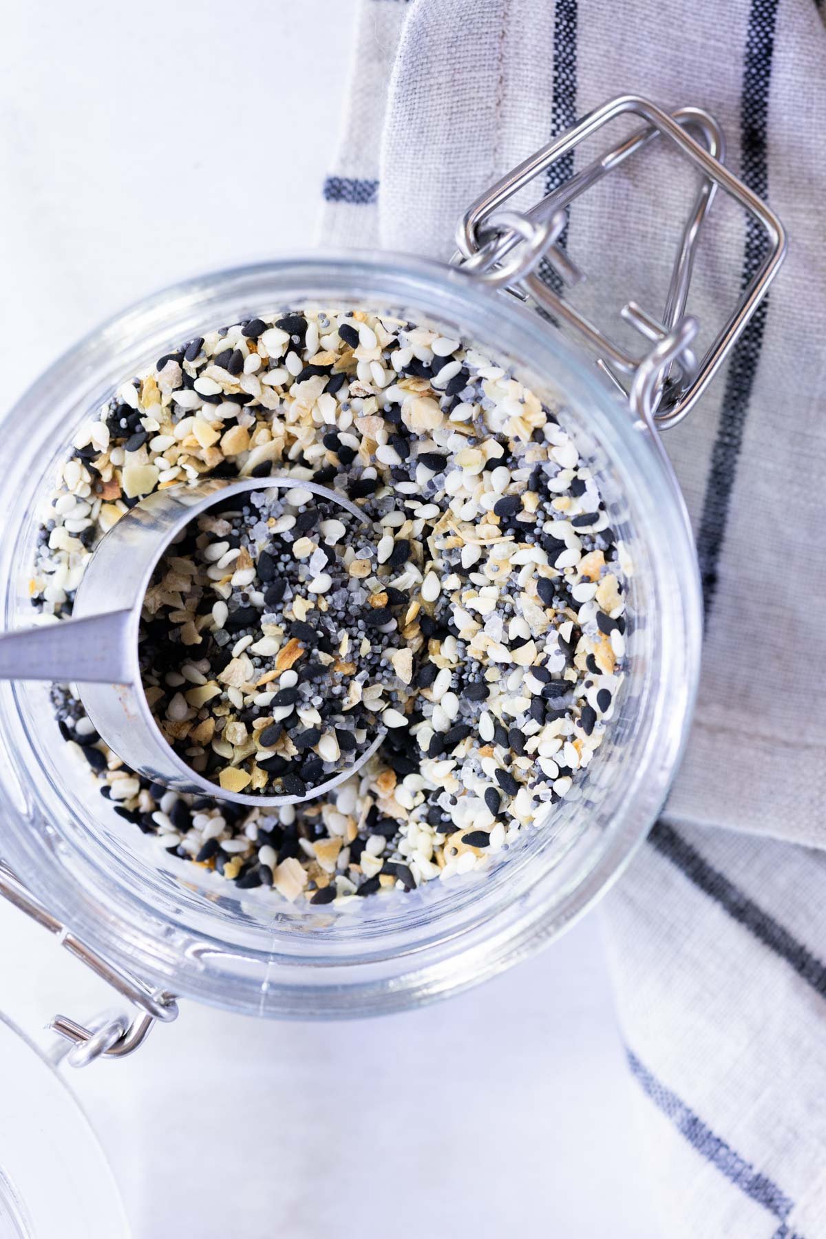 Glass jar filled with everything bagel seasoning mix and a metal scoop, placed on a light surface next to a striped cloth napkin.